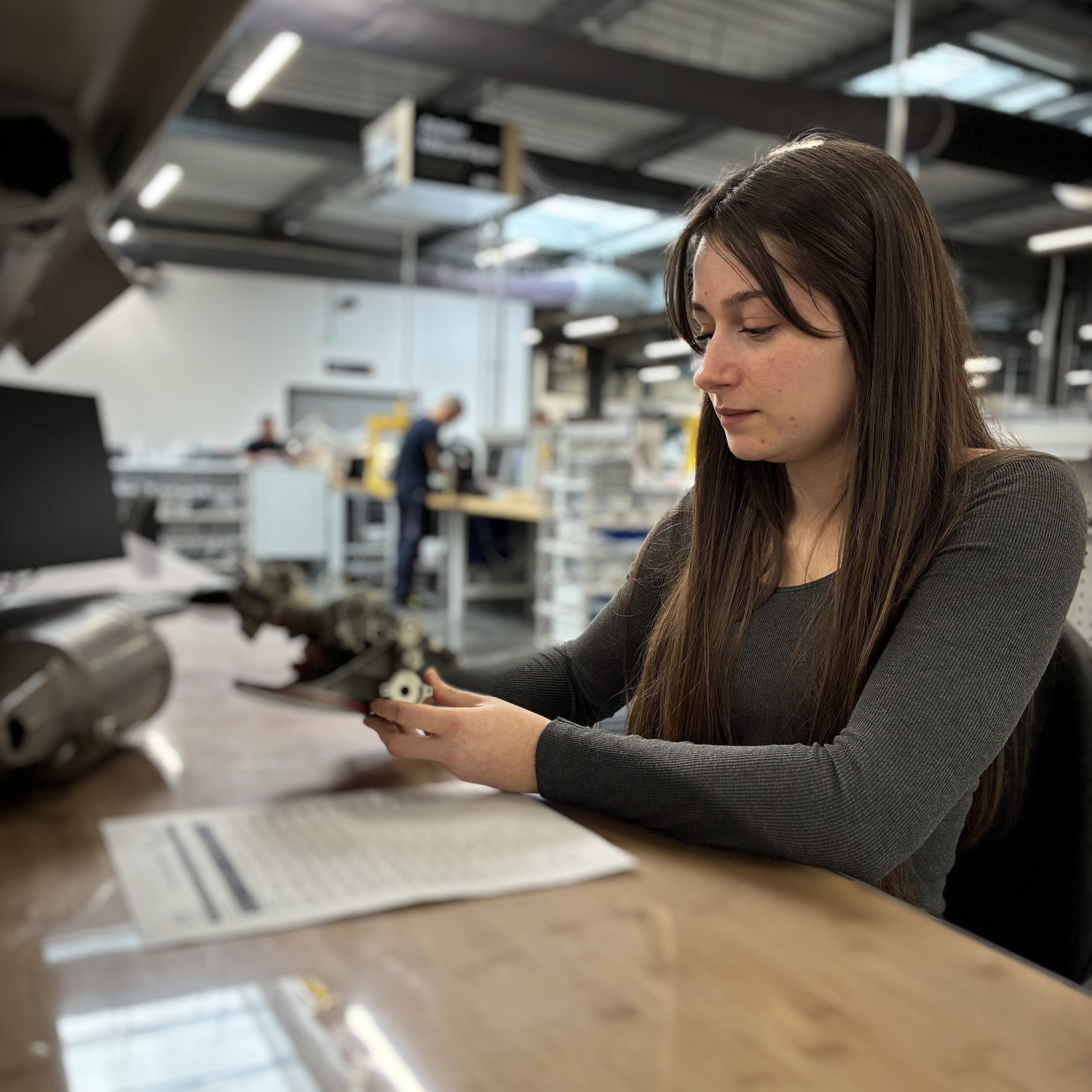 A young woman with long hair sits at a wooden workbench in a large industrial workshop, carefully examining a small mechanical part she holds in her hands. A paper document lies on the table in front of her. In the background, the vast workshop, with its industrial ceiling structure, workstations, and other professionals at work, creates a subtle, artistic blur.