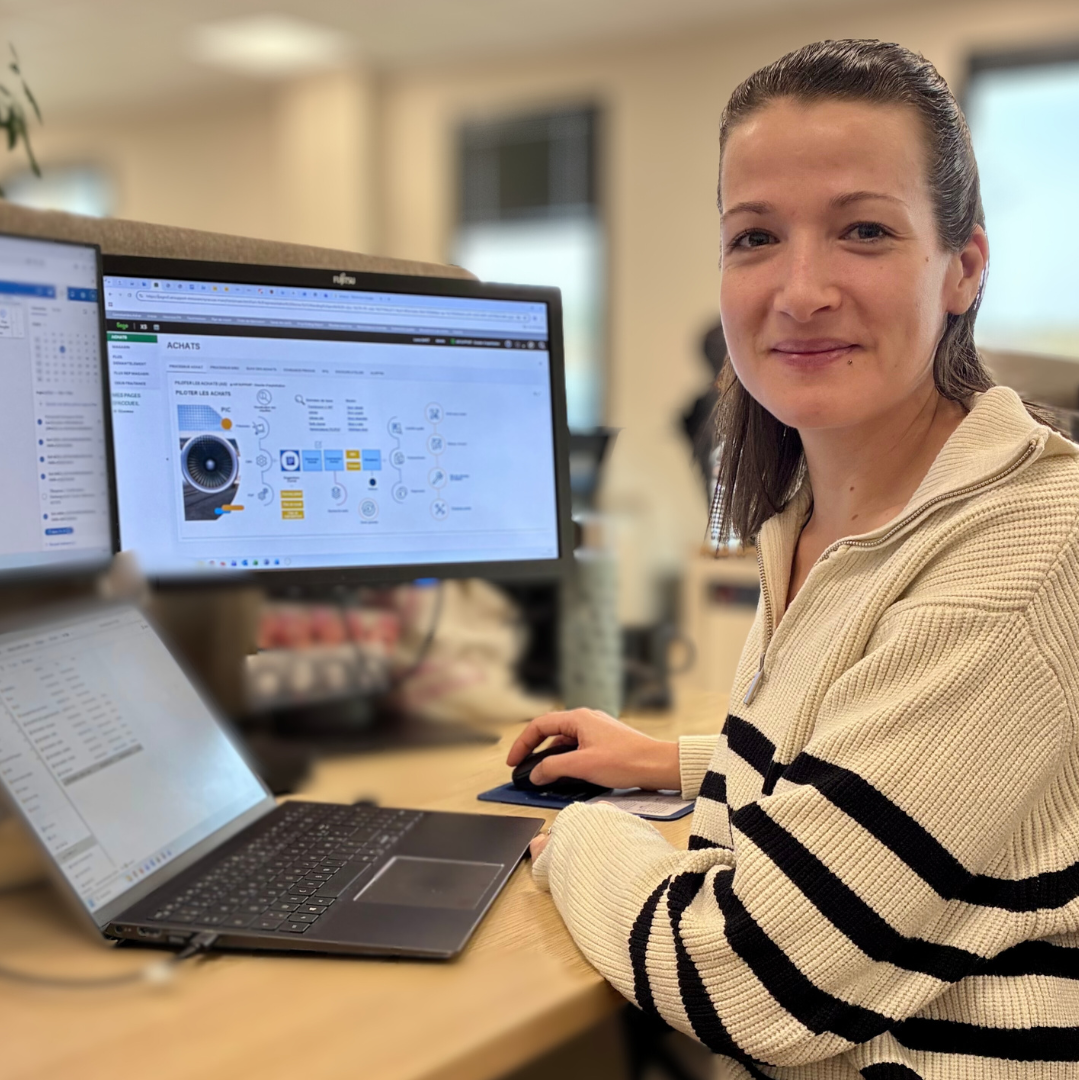 A smiling woman, wearing a black and white striped sweater, sits at her desk and looks at the camera. Her hand rests on a computer mouse. In front of her are an open laptop and two large computer monitors. The main monitor displays professional management software called 
